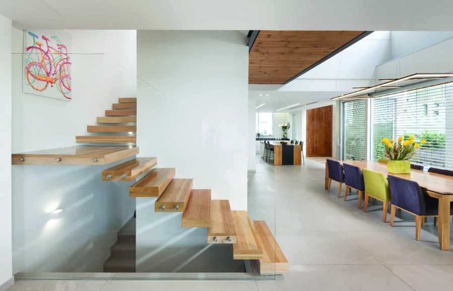 מבט מעומק חלל המגורים לכיוון פינת האוכל, המטבח וגרם המדרגות המרחף שעוטף את גרעין המעלית ומוביל אל הקומה העליונה.