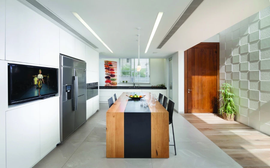 המטבח שבקדמת מרחב המגורים: יחידת קיר גדולה, משטח כיור/עבודה עם חיפוי זכוכית מודפס, ופסי תאורה בתקרה שמדגישים את הקו הליניארי של חללו. יחידת אי מרכזית מעוצבת מעץ אלון עם 