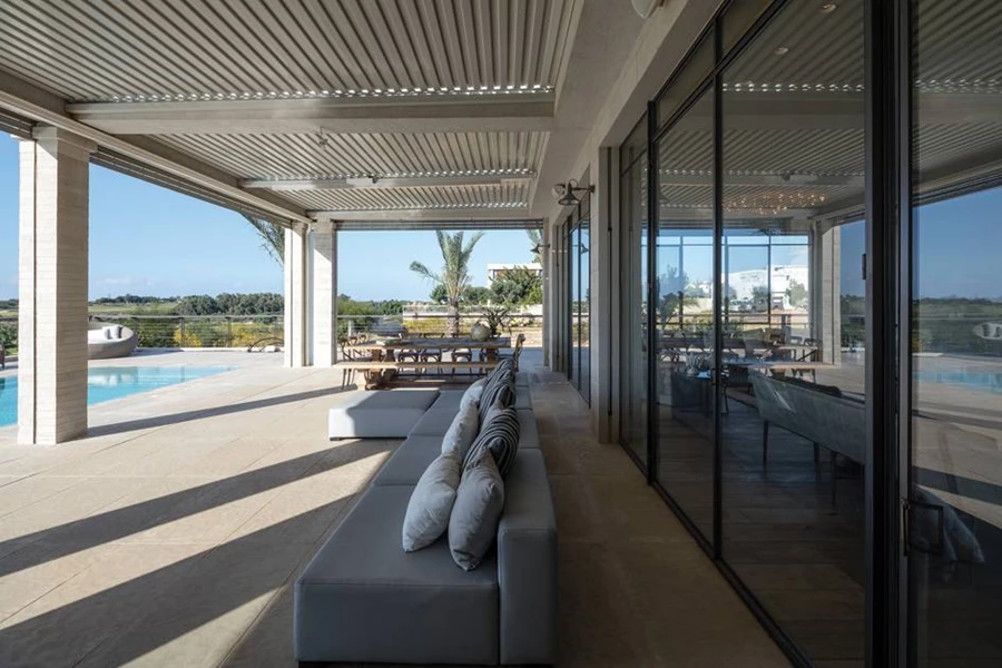 מערך ויטרינות המייצר חזית שקופה לחלל המגורים חושף את רחבת הישיבה החיצונית. הרחבה מקורה בתריסים חשמליים הניתנים לסגירה מלאה ומאפשרים להפכה לאזור נוסף של חלל הפנים.