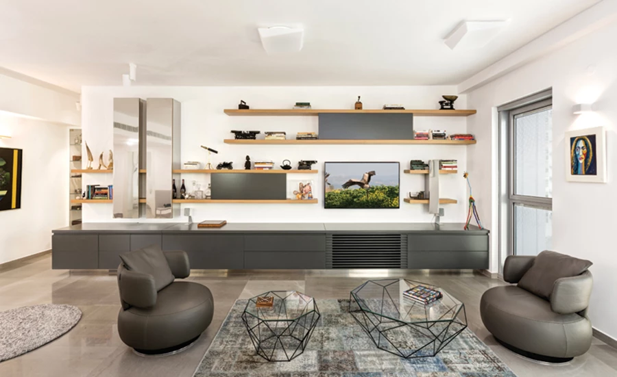 לאורך קיר הסלון: קומפוזיציה המשלבת מדפים פתוחים מעץ אלון, תאים סגורים, שתי יחידות אנכיות עם חזיתות נירוסטה בגימור מראה שמשמשות כבר, ויחידת אחסון נמוכה הנמשכת לכל אורכה ומכילה, בין היתר, מקרר יינות. ממול: ניגוד צורני בין שתי כורסאות בעיצוב מעוגל לבין שולחנות קפה בעיצוב אוורירי מצולע. עיצוב וביצוע ספרייה: יפרח בן צבי, מערכת ישיבה: פיטרו הכט, תאורה: קמחי תאורה, שטיחים: צמר שטיחים יפים.