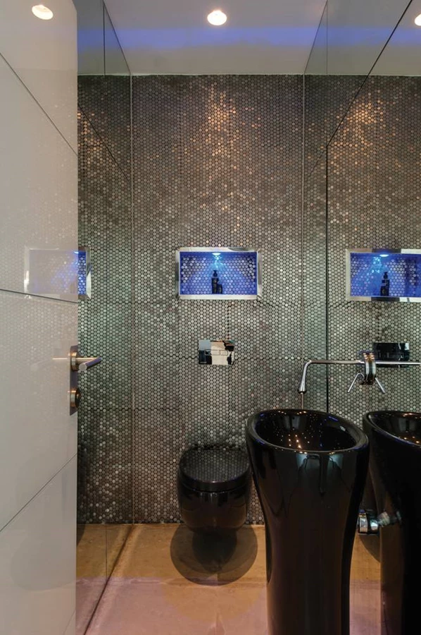 שירותי האורחים: עיצוב דרמטי המשלב חיפוי קיר באריחי פסיפס עשויים נירוסטה, גומחה קטנה מוארת באור לד כחול, ואביזרים סניטאריים מסוגננים בגון שחור.