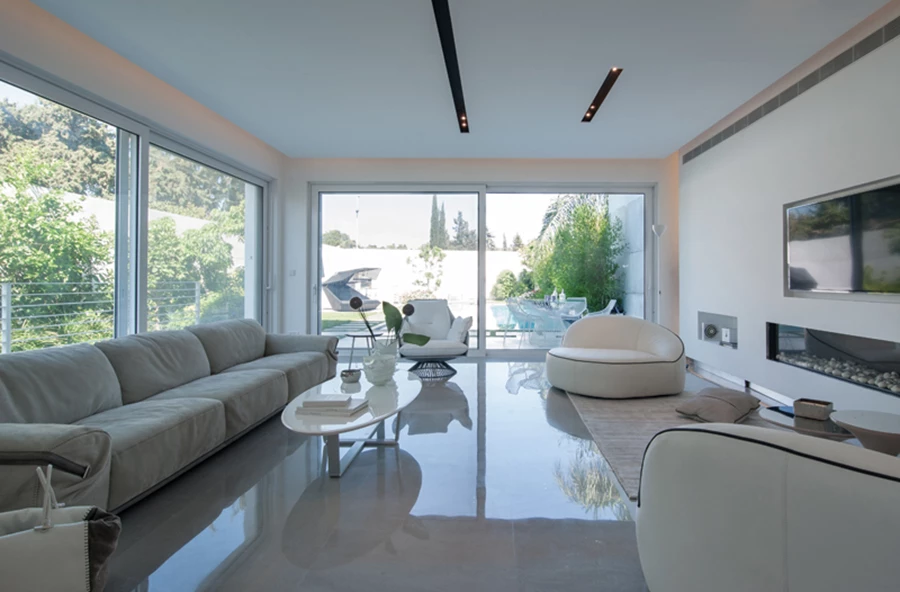 אזור הסלון: עיצוב מינימליסטי, בהיר ומזמין. ריצוף באריחי שיגריי אפורים בגימור מלוטש, תאורה שקועה וסמויה, וויטרינות הקושרות אותו ישירות אל החוץ. ריהוט: ״אליתה ליוינג