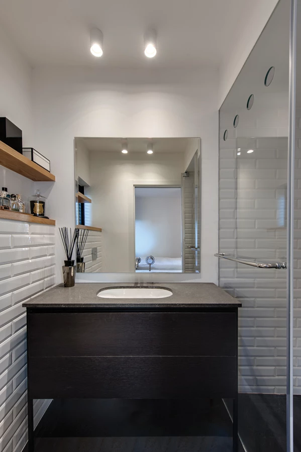 חדר הרחצה: חיפוי קיר באריחים לבנים המייצרים מופע גראפי, ויחידת כיור מעוצבת מסגרת ורגליים מברזל, מגירות פורניר אלון מושחר ומשטח בזלטינה.