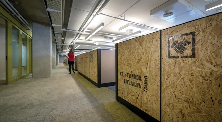 חלוקת החלל החדשה: שדרת מעבר מפרידה בין אזור העבודה המשותף לבין משרדי המנהלים, מלווה במקצב עמודי הבטון התומכים שנותרו בחלל ומחופה בשטיח בגווני חרדל משתנים, כעין התזת ספריי לא אחידה. מחיצות חדרי מנהלים ושטיחים מבית interface -