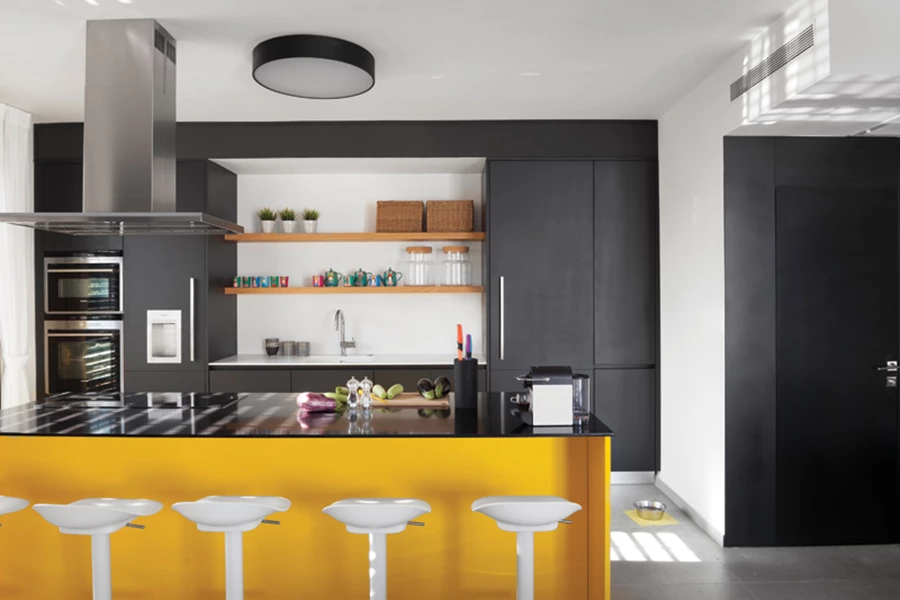 המטבח: יחידת קיר באפור גרפיט הכוללת בתוכה מעין גומחה בה משתלבים מדפים פתוחים, משטח עבודה וכיור. יחידת האי בצהוב עז עם משטח אבן קיסר שחור. שירותי האורחים הסמוכים עוצבו בהתאמה עם דלת שעוצבה ייעודית בנגרות זהה ובקו אפס עם הקיר. מטבח: