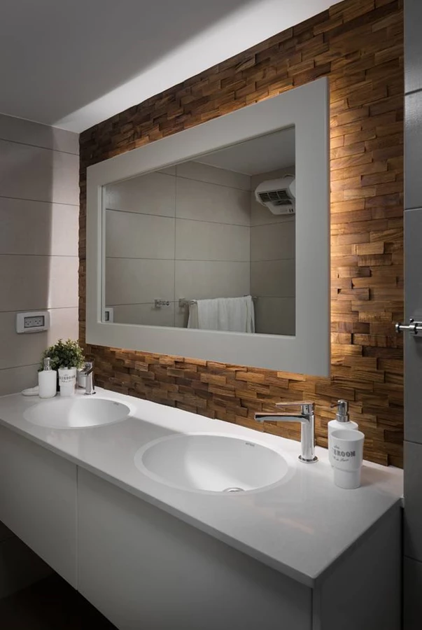 חדר הרחצה של הבנות: משטח כיור עם כיורים אינטגראליים, חיפוי קיר תלת ממדי באריחי עץ, והילת אור הבוקעת סביב המראה המותקנת מעליו.