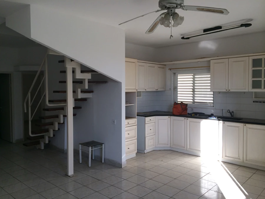 מבט מהסלון אל עבר המטבח הפתוח וגרם המדרגות: יחידות מרחפות עם חזיתות פורמייקה במראה מתכתי, ונקודת חן בדמות מדפים אדומים תלויים על שרשראות.