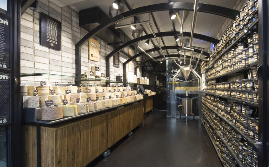 החלבה מול אריזות הטחינה- לכל מוצר הוקדשה דופן, ומעל- צנרת תעשייתית שמובילה את גרגירי השומשום אל אבני הריחיים. חיפוי קירות: בריקים רטרו בגווני אפור ״קרן סטון״, תאורת לד מקצועית: דורי קמחי תאורה״.