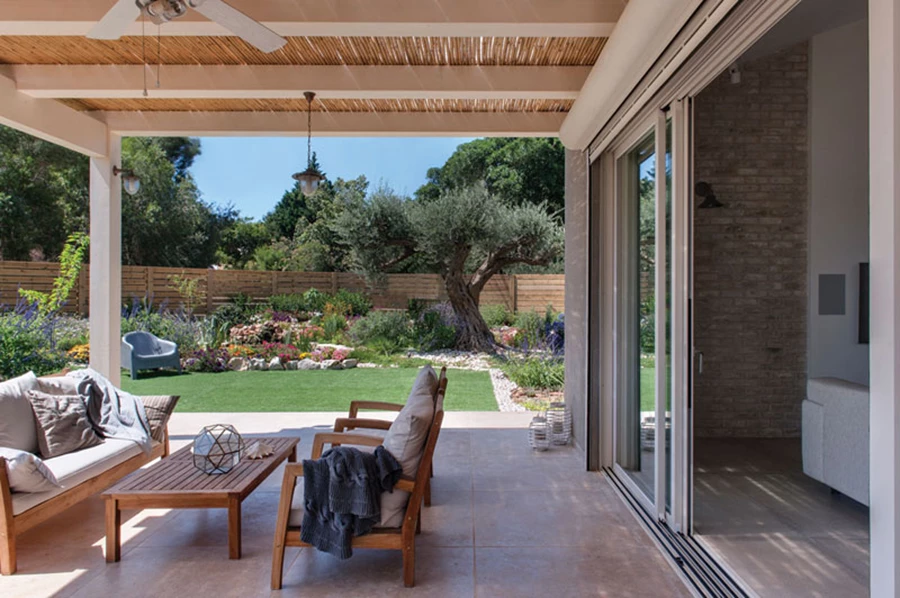 ביציאה מחדר המגורים: פרגולה לבנה מקרה רחבה מרוצפת באבן סאני מכובסת המאכלסת בשטחה פינות ישיבה ואוכל המשקיפות אל מרחב הגן המטופח. ריצוף רחבת ישיבה: