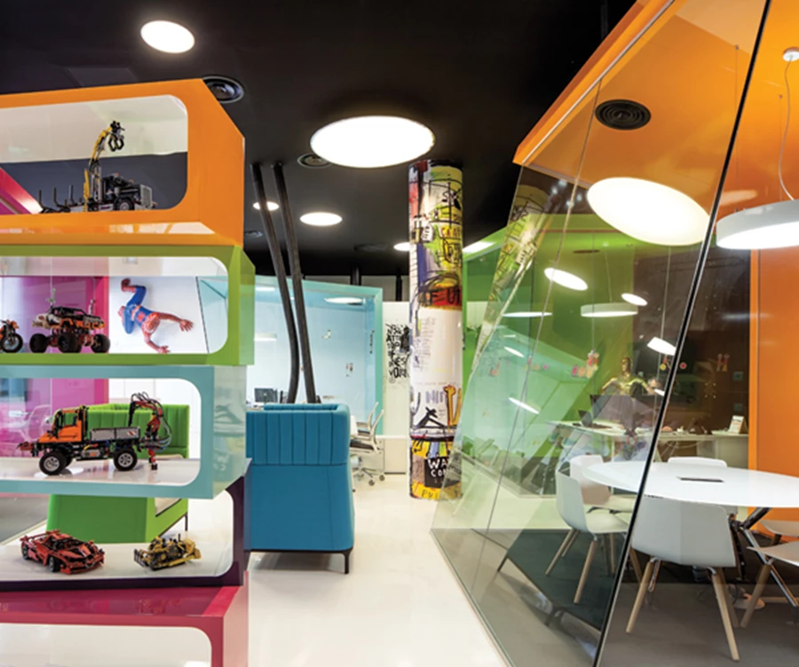 מבט מהמעבר אל עבר חלל המשרד: ספרייה צבעונית מעוצבת כעין ערמת תיבות הסדורות בהזחה זו מעל זו  תוחמת אותו כלפי המרחב הפתוח. צבעוניות הפביליונים נגזרה מצבעי התיבות.