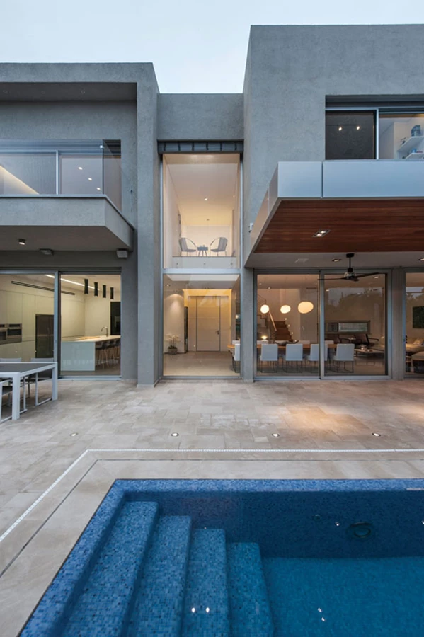 מבט מעומק הגינה אל עבר הבית חושף את קומת הקרקע, את החלל הגבוה הסמוך לויטרינות ואת פינת הישיבה הקטנה בגשר הקומה העליונה. ריצוף הרחבה באריחי אבן קרמה מוס בעיבוד מוברש וסידור פטרן.