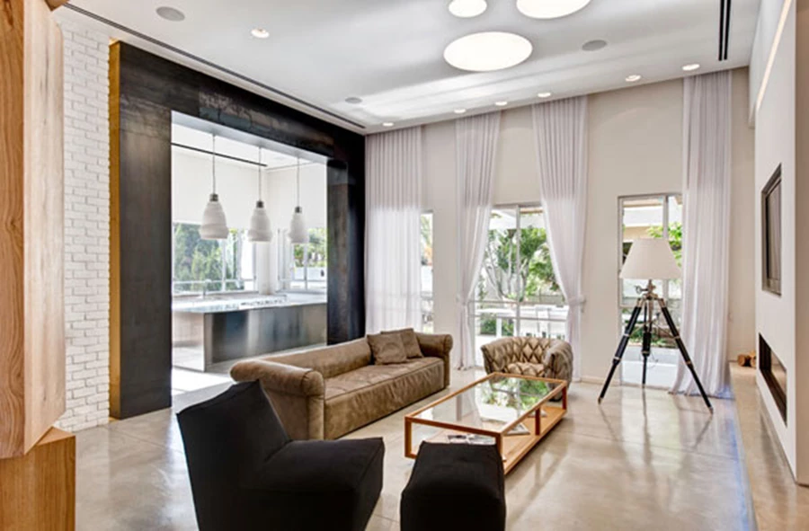 אזור הסלון: עיצוב נקי ומדוד, וילונות אווריריים, ושער ברזל גבוה המדגיש את המעבר למטבח. אח אינטגראלי ומסך טלוויזיה מוטמעים בתוך אלמנט גבס מעל ספסל בטון מרחף עם תאורה תחתיו. קורות חיפוי ברזל בשער המטבח: