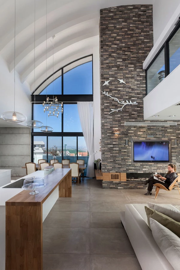 מבט מהכניסה לבית: מרחב שמרביתו הינו חלל גבוה תחום בפתחים גדולים החושפים את הנוף כשצורתם נגזרת מהגג המעוגל. מימין נראית קומת הגלריה העליונה, וקיר מחופה לבנים מגדיר את אזור הסלון במרחב הפתוח. ריצוף: אריחי גרניט פורצלן המדמים מראה של בטון. ריצוף: