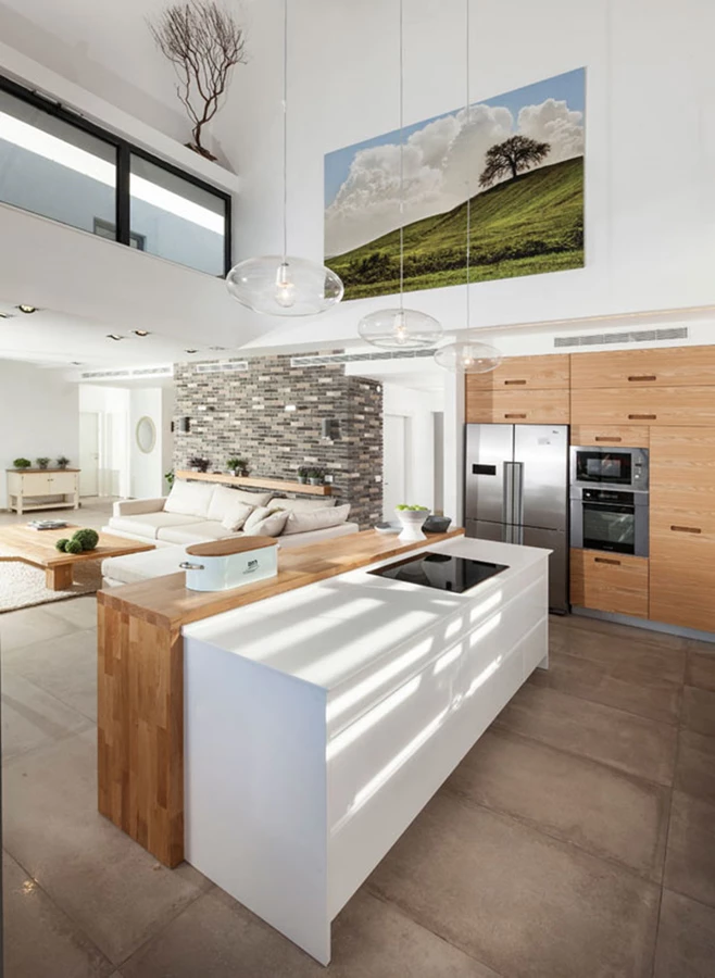 מבט מהמטבח אל עבר הגלריה העליונה, הסלון, והחלל המצוי מאחוריו בו ממוקמים יחידת הורים וחדר נוסף. אל יחידת הקוריאן הלבנה חוברת יחידה קונזולית עשויה עץ אלון צרפתי, ובחלקו העליון של הקור נראים הצילומים ששולבו בבית.