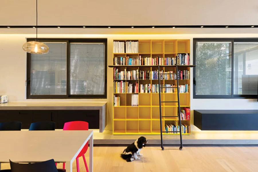 מול המבואה, במעבר חד מהדרמה הכהה: ספרייה בצהוב עז ושתי יחידות נגרות המרחפות מעל משטח בטון תחתון. לריצוף החלל משמש פרקט בהיר. פרקט: