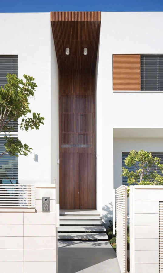 תמונה קטנה-שמאל-חזיתו הקדמית של הבית: מדרגות שיש מרחפות ומרחב כניסה חיצוני שגובהו מתנשא עד גובה הגג. חיפוי סרגלי עץ האלון המגוון של הדלת ממשיך מעלה ועוטף גם את הקירוי, כשהוא מותאם לסרגלי עץ הטיק של חלונות הקומה השנייה.