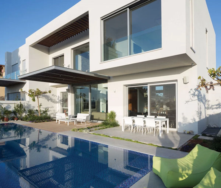 מבט מעומק הגינה אל חזיתו האחורית של הבית. רחבת ישיבה מרוצפת באריחי שייגריי זהים לאלה שבבית, דק עץ עם כורסאות מזמינות, ובריכת גלישה מחופה באריחי פסיפס כחולים.