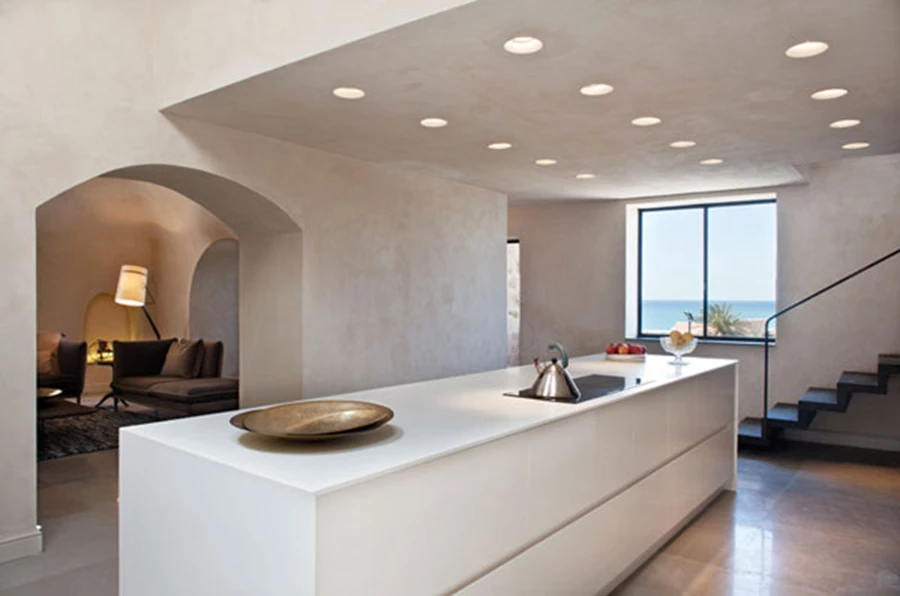 מבט אל המטבח הלבן המעוצב בשפה מודרנית-מינימליסטית. ריצוף באריחי שייגריי אפורים, חיפוי קירות בטיח מינראלי, ופתחי סקיילייט בתקרה שנפרצו כנראה במסגרת אחת מהרחבות הבנייה שעבר החלל.