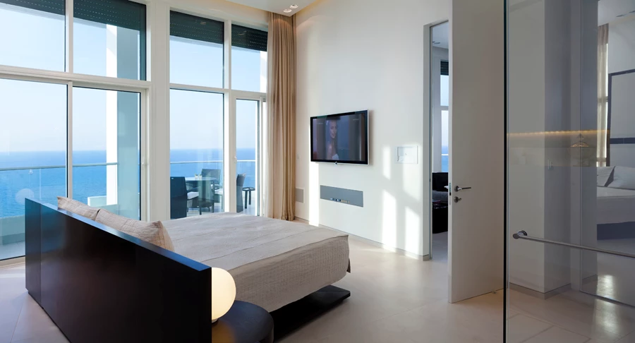 יציאה למרפסת גם מחדר השינה. האלומיניום בויטרינות נצבע בגון לבן והארון שמאחורי המיטה משתלב בתוך גומחה בקיר.