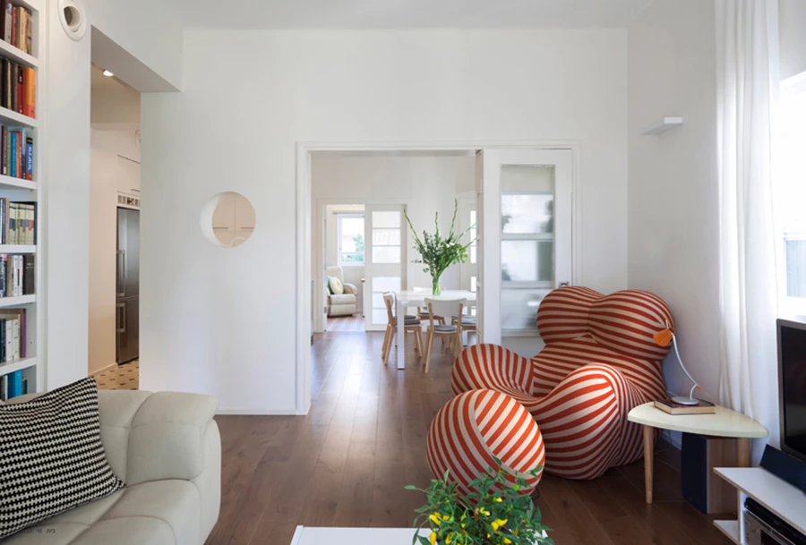 ציר קשר ששומר בתכנון החדש: חלל הסלון מקושר באמצעות דלת כנפיים מקורית ששופצה בתהליך רסטורציה. קירות לבנים, ריצוף בפרקט עץ אלון, ופתח עגול מייצר קשר עין עם חלל המטבח. בעומק נראה חדר העבודה ש