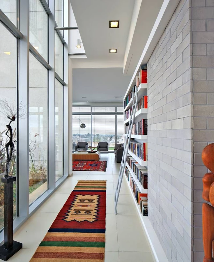 המבואה הפנימית: מוגדרת באמצעות אלמנט הספרייה המחולל את התנועה בבית. זורמת כציר אורכי ומושכת את המבט לכיוון הסלון. חיפוי לבני סיליקט המשתלב לצד המדפים: תוצר של עומק היחידה שגויס לטובת שילוב המקרר במטבח הממוקם מאחוריה. נגרות ספרייה: