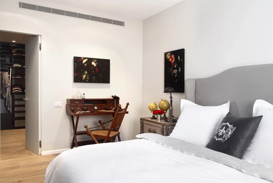 חדר השינה המרווח נחלק לאזורים ומעוצב בשפה אקלקטית, מובחנת ואנינה. כאן נראה אזור השינה בו משתלבים מיטה מרופדת עם גב גבוה עשיר למראה, סקרטר קלאסי בטעם של פעם, ויצירות אמנות.