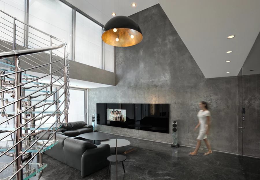 מבט מהכניסה אל חלל המגורים: חלל כהה, גבוה ועוצמתי, שבמרכזו ניצב גרם מדרגות מעוגל עשוי נירוסטה ומדרכי זכוכית. ריצוף באריחי שיש כהים עם משחקי גוונים וגידים, קירות ב