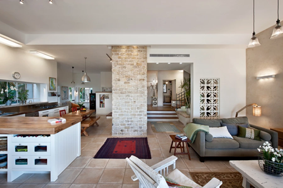 מבט מכיוון עומק החלל אל עבר מבואת הכניסה: הסרת הקירות שתחמו את המטבח יצרה תחושת מרחב וזרימה פתוחה בין אזורי החלל השונים. חלקת קיר עם משרבייה המשתלבת בתוכה מצניעה מאחוריה את הירידה למרתף. משרבייה: 