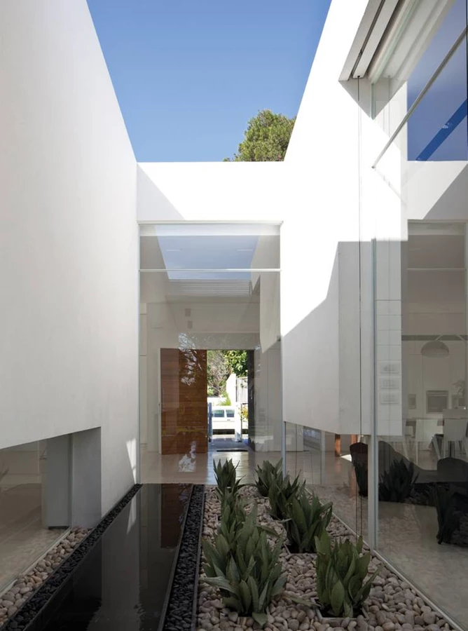 מבט מהגן המשתרע בחזית האחורית אל עבר בריכת השיקוף ודלת הכניסה: קונספט רעיוני הנשען על ציר אורכי הכולל פטיו עם בריכת שיקוף, אשר למרות היותם חיצוניים מהווים חלק בלתי נפרד מחלל הפנים.