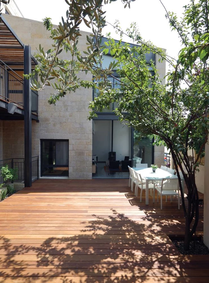 החצר הפנימית במבט הגשר - תחומה בין צלעות ה-L של הבית ובקירות תומכים, כשהיא מוצנעת מעין העוברים ושבים ברחוב ומבטיחה פרטיות ונוף עצים ירוק. קיר שקוף קושר אותה פיזית וויזואלית לחלל הפנים, ושטחה המרוצף עץ איפאה מאובזר כראוי להנעמת השהייה בה