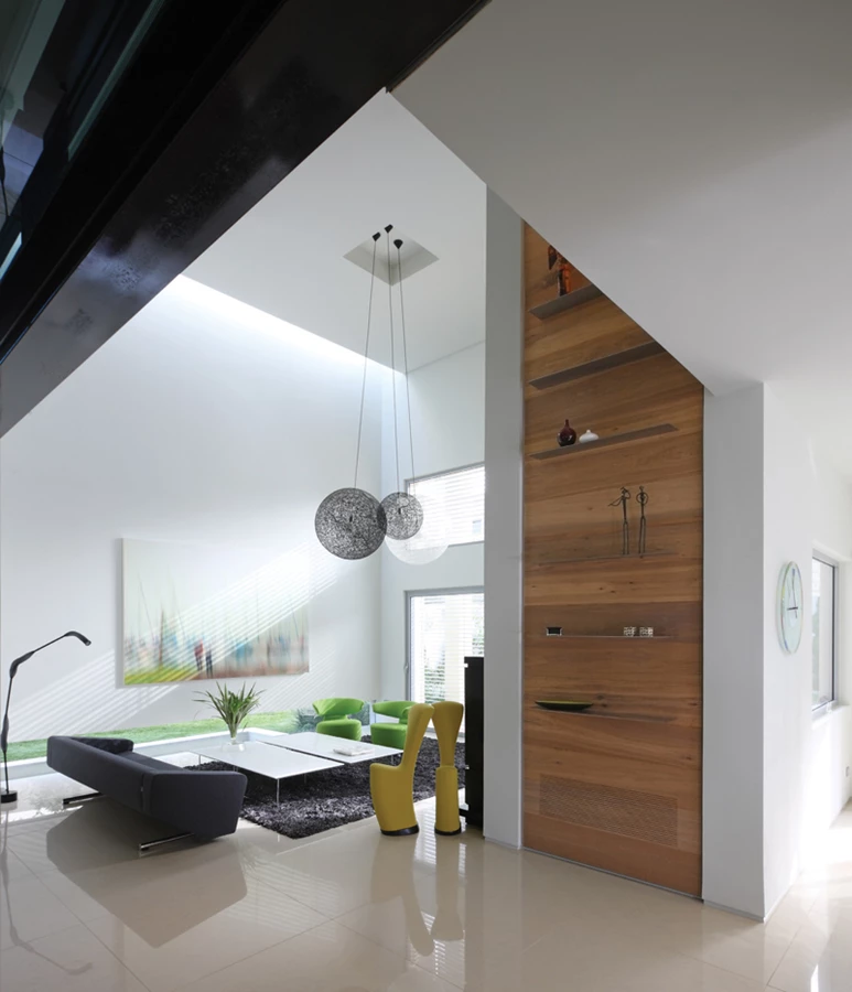 מראה מדלת הכניסה - קונטור המעטפת מייצר את הגדרת אזורי החלל, ללא תוספת של מחיצות בנויות נוספות. הקיר המפריד בין אזורי המטבח והסלון מחופה בלוחות פרקט אלון, ועליו מותקנים מדפי מתכת מעודנים להצבת פריטי נוי מוקפדים. ריצוף באריחי גרניט פורצלן בהירים בגימור מבריק מדגישים תחושת ניקיון. כורסאות צהובות: “הביטאט”.