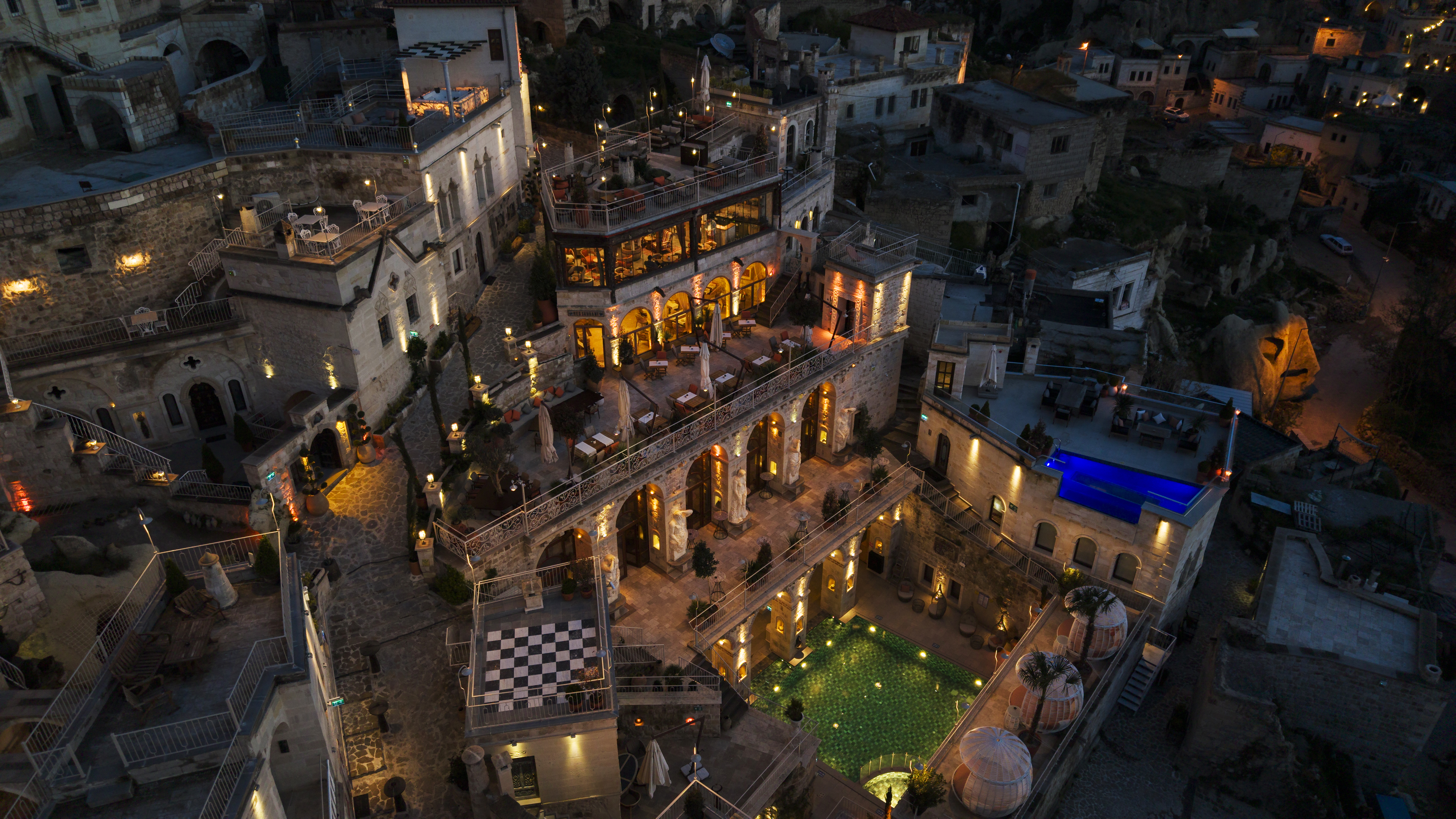 מתחם האבן של Elika Cave Suites Cappadocia בלילה, עם חצר שבה בריכה מרוצפת בירוק ובריכה מוארת בכחול על הגג. © 2026 Hilton