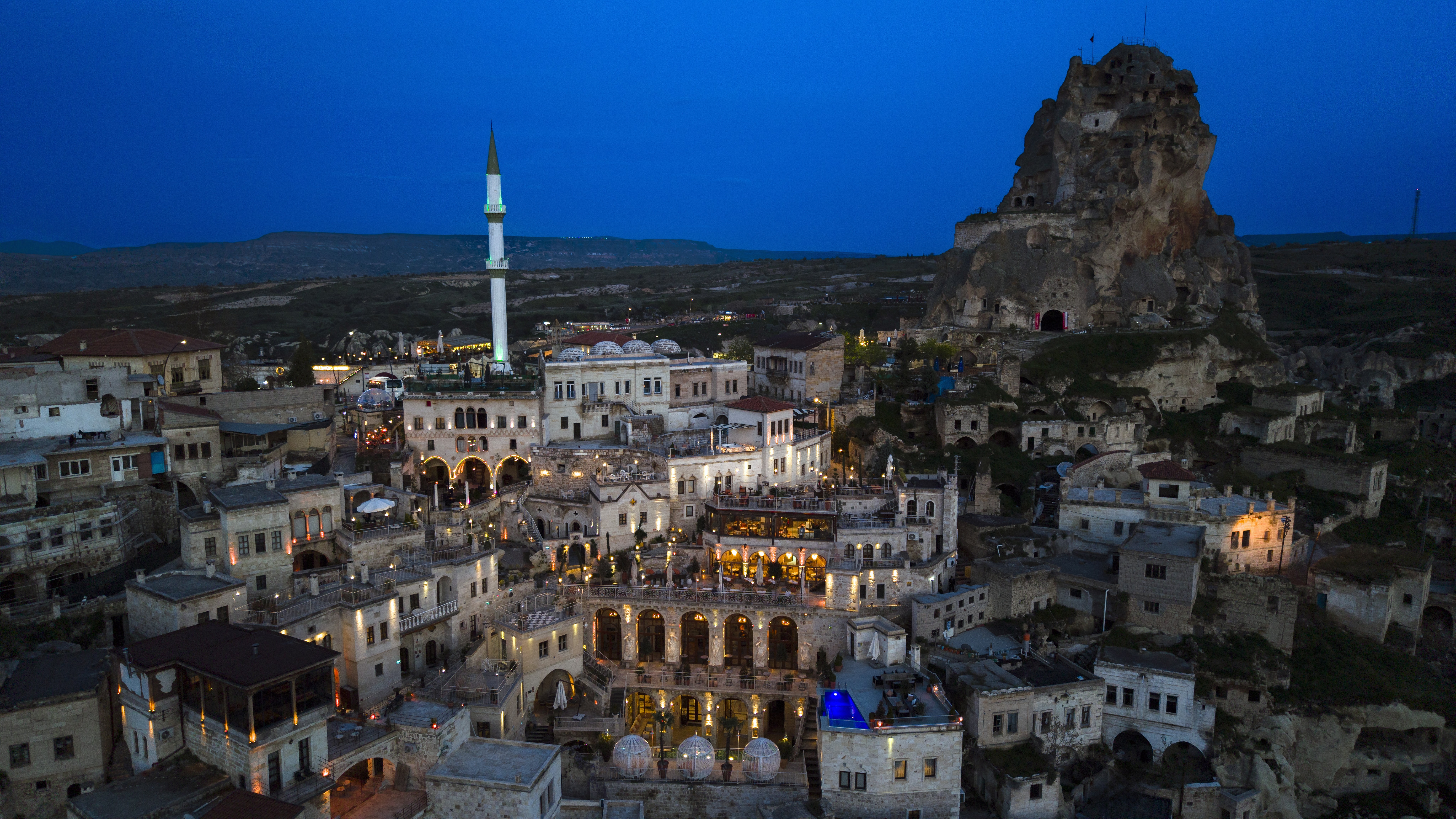 מבני האבן של Elika Cave Suites Cappadocia באורטהיסאר, עם חלונות מוארים, מינרט וסלע גדול ברקע. © 2026 Hilton
