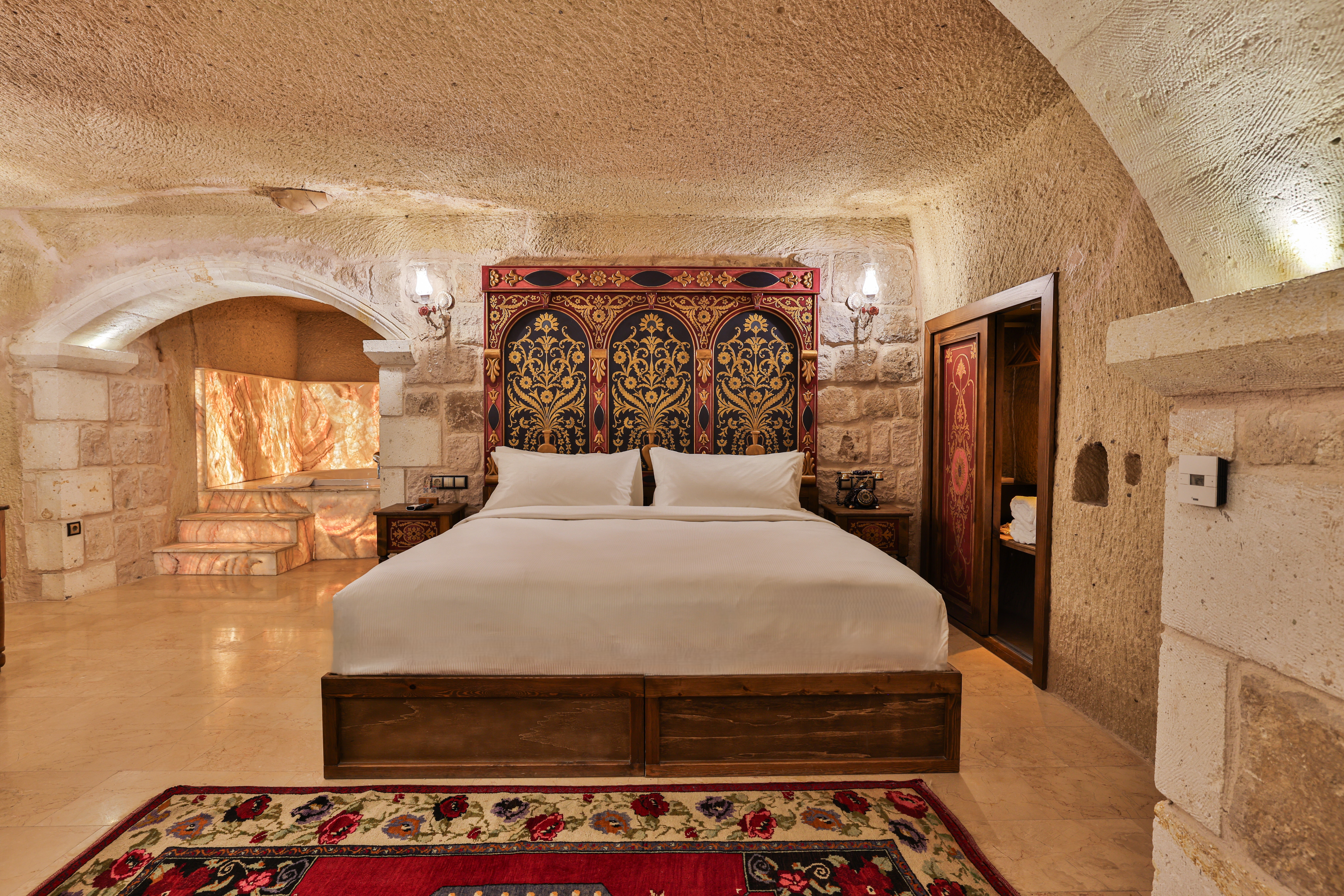 חדר King Grand Cave ב־Elika Cave Suites Cappadocia, עם קירות אבן, מיטה גדולה עם בסיס מעוטר ושטיח פרחוני צבעוני. צילום © 2026 Hilton