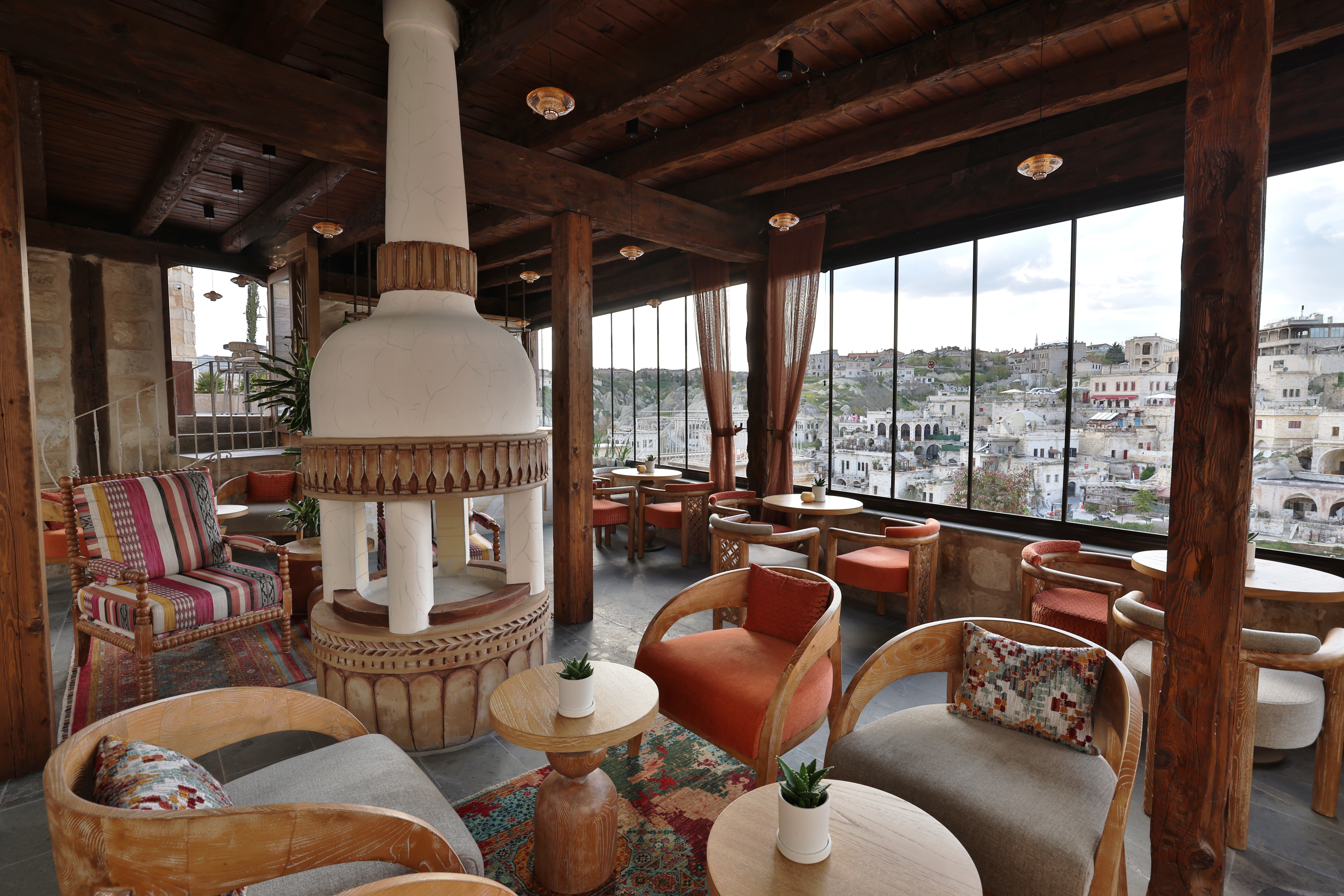 ה־Colastre Lounge Bar ב־Elika Cave Suites Cappadocia, עם פינת ישיבה, כיסאות עץ, כריות צבעוניות ואח לבן במרכז החלל. החלונות הגדולים משקיפים על עיירה על צלע גבעה. © 2026 Hilton